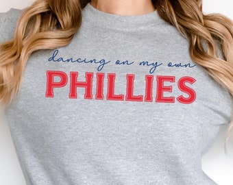 Może przedstawiać: Szara bluza z napisem "dancing on my own PHILLIES" w czerwonych i niebieskich literach.