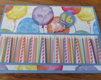 May include: A handmade birthday card with a blue background and a watercolor design of balloons and candles. The card has a blue circle with the number 16 in yellow.