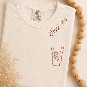 May include: An off-white short-sleeve t-shirt featuring brown embroidery on the chest. The text reads "Hook em" in a cursive script above an embroidered hand gesture showing the "hook 'em horns" sign. The shirt has a Comfort Colors brand label.