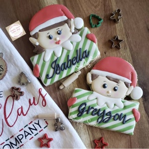 May include: Two elf-themed cookies, one labeled "Isabella" and the other "Greyson", with green and white striped rectangles. Both elves wear red hats. Cookie cutters and a rolling pin are scattered around.