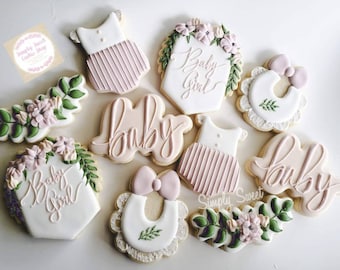 May include: A set of nine decorated sugar cookies with pink and green frosting. The cookies feature various baby-themed designs, including a bib, a onesie, and the words "baby" and "baby girl".