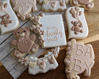 May include: Assortment of decorated sugar cookies in a baby shower theme. Cookies include teddy bears, balloons, baby clothes, and the words "We can bearly wait" and "BABY". The cookies are decorated with brown, white, and beige icing.