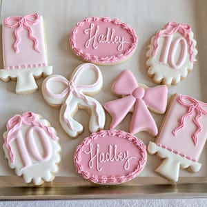 May include: A collection of decorated sugar cookies in a baking pan. The cookies are primarily pink and white, featuring bow and cake designs, the number 10, and the name "Hadley" in cursive script. The cookies are arranged on a light-colored surface.