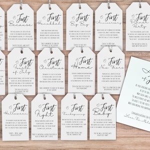 May include: A set of 14 white gift tags with black calligraphy. Each tag features a heart and the text "First" followed by a special occasion, such as "First Big Trip" or "First Christmas". The tags are arranged in two rows on a wooden surface. A white card with black calligraphy reads "A Year of Firsts" and includes a short message about celebrating milestones.