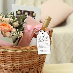 May include: A wicker basket filled with a bouquet of peach roses, a box of chocolates, a bottle of wine, and a gift tag that reads "First Baby".