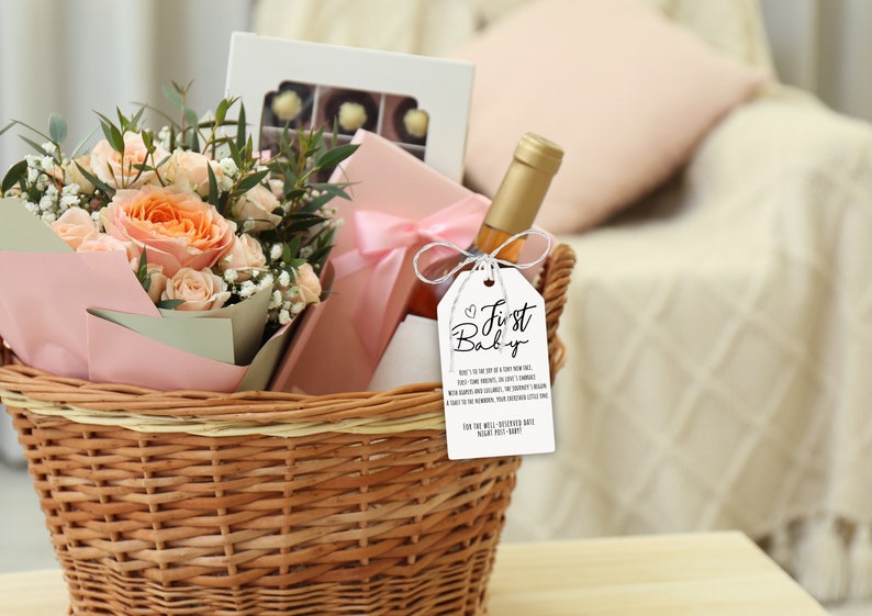 May include: A wicker basket filled with a bouquet of peach roses, a box of chocolates, a bottle of wine, and a gift tag that reads "First Baby".