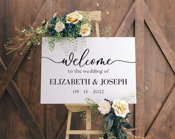 May include: White wedding welcome sign with black calligraphy text that reads "Welcome to the wedding of Elizabeth & Joseph 09-15-2022". The sign is propped up on an easel and decorated with white and yellow flowers.