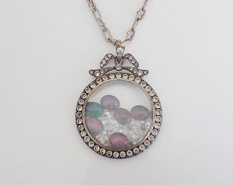 May include: A silver locket necklace with a round, clear glass window. The locket is decorated with a bow and small, clear stones. Inside the locket are several small, colorful stones.