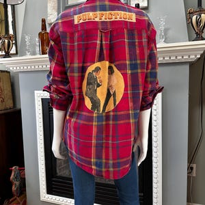May include: Red plaid button-down shirt with the text "PULP FICTION" across the shoulders. The back of the shirt features a circular graphic with two figures. The shirt is paired with blue jeans.