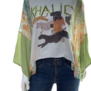 May include: White graphic tee with the name "KHALID" in green letters, featuring a colorful illustration of a person and animals. The tee is paired with a patterned, flowing overlay and dark blue jeans.