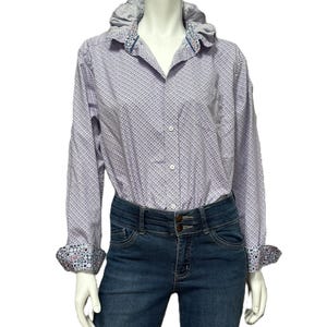 May include: A light purple button-down shirt with a small geometric pattern, paired with dark blue denim jeans. The shirt features a ruffled collar and patterned cuffs. The jeans have a double-button closure.