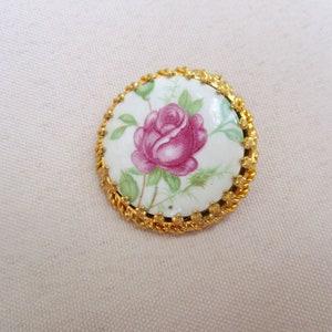 Könnte beinhalten: Eine runde, goldfarbene Brosche mit einem weißen Porzellanmittelteil, das eine rosa Rose mit grünen Blättern zeigt.