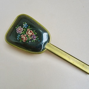 Könnte beinhalten: Ein goldfarbener Handspiegel mit einem schwarzen Blumendesign. Der Spiegel ist oval geformt und hat einen dekorativen Griff.