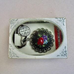 Könnte beinhalten: Eine schwarze und silberne Halskette mit einem runden Anhänger, der mit einem floralen Stickmuster verziert ist. Der Anhänger ist von einem silbernen Filigranrahmen umgeben. Die Halskette ist in einer weißen Schachtel verpackt, auf der "Handarbeit" und "Rot-Print" steht. Die Schachtel trägt außerdem den Schriftzug "Made in Austria".