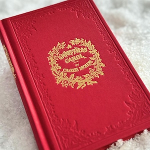 May include: A red hardcover book titled "A Christmas Carol by Charles Dickens." The book has gold lettering and a decorative wreath design. The cover has embossed floral corner accents.