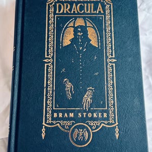 May include: A hardcover book titled "DRACULA" by Bram Stoker. The cover is dark blue with gold lettering and illustrations, including a silhouette of a figure and a bat emblem. The book is closed and resting on a white surface.