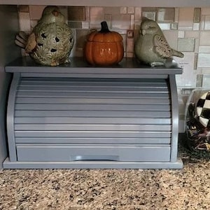 May include: A gray roll-top bread box with a wood-like finish sits on a countertop. The bread box has a decorative roll-top lid and is accented with two ceramic bird figurines and a small pumpkin.