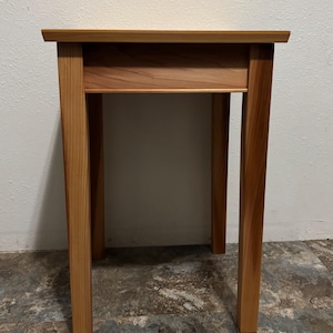 May include: A small, rectangular wooden side table with a natural finish. The table has a simple design with four straight legs and a single shelf. The wood grain is visible, and the table appears to be sturdy. The table is approximately 30 inches tall.
