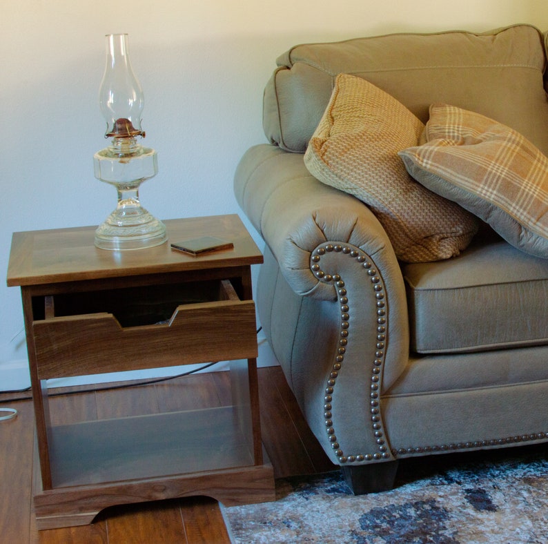 Puede incluir: Una mesa auxiliar de madera con un caj&oacute;n y una l&aacute;mpara de aceite de vidrio en la parte superior. La mesa est&aacute; junto a un sof&aacute; gris con dos almohadas.