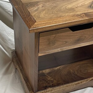 Puede incluir: Una mesa auxiliar de madera marr&oacute;n oscuro con un caj&oacute;n y un estante. La mesa tiene un borde decorativo en la parte superior e inferior.