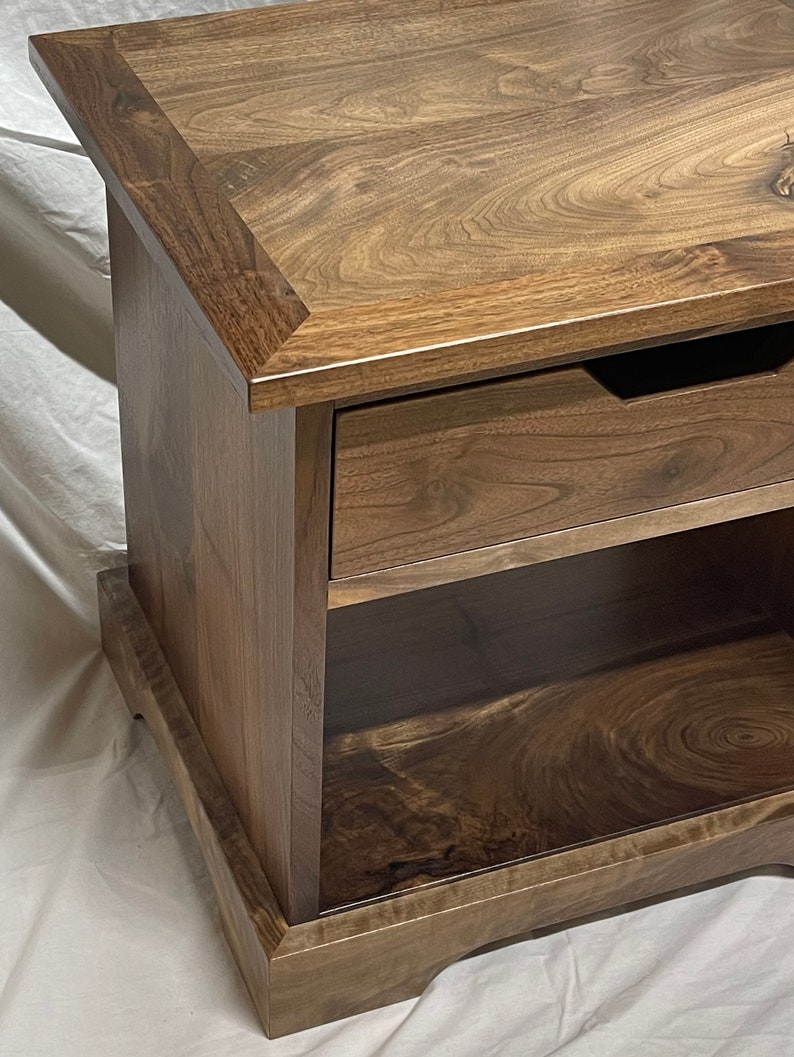 Puede incluir: Una mesa auxiliar de madera marr&oacute;n oscuro con un caj&oacute;n y un estante. La mesa tiene un borde decorativo en la parte superior e inferior.