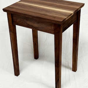 Puede incluir: Una mesa auxiliar de madera hecha a mano con una parte superior cuadrada y cuatro patas rectas. La mesa está hecha de madera marrón oscuro con un patrón de vetas natural. La mesa mide aproximadamente 61 cm de alto.