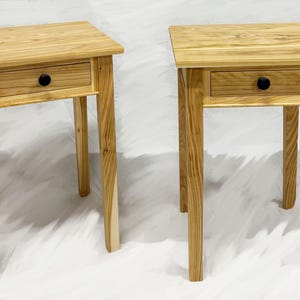 May include: Two wooden side tables with a natural finish. Each table has a square top, a single drawer with a black knob, and four legs. The tables are made of light-colored wood and are set against a white background.