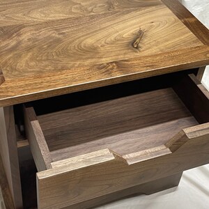 Puede incluir: Una mesita de noche de madera con un solo caj&oacute;n. El caj&oacute;n est&aacute; abierto y se ve la veta de la madera. La mesita de noche est&aacute; hecha de madera oscura con un acabado marr&oacute;n claro.