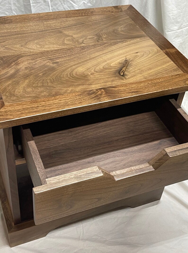 Puede incluir: Una mesita de noche de madera con un solo caj&oacute;n. El caj&oacute;n est&aacute; abierto y se ve la veta de la madera. La mesita de noche est&aacute; hecha de madera oscura con un acabado marr&oacute;n claro.
