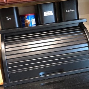 May include: Black bread box with a roll-top lid. The bread box is sitting on a counter with three black canisters labeled "Tea", "Decaf Coffee", and "Coffee".