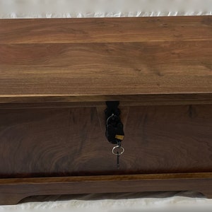 May include: A dark brown wooden storage chest with a keyhole and a key in the lock. The chest has a raised edge around the top and a base with a slight curve.