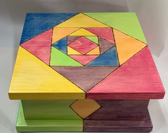 Puede incluir: Una caja de madera con un diseño geométrico pintado en tonos de amarillo, naranja, rosa, verde, azul y morado. La caja tiene una tapa con bisagras y está hecha de madera.