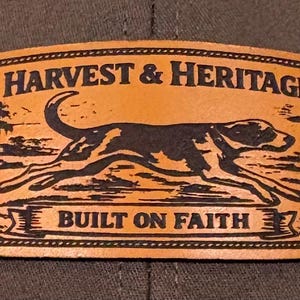 May include: A leather patch with the words "HARVEST & HERITAGE" and "BUILT ON FAITH" in a rectangular shape. A black dog is running across the center of the patch. The patch is orange with black text and a black border.