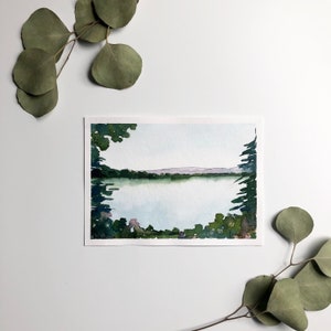 May include: Watercolor painting of a serene lake scene. The artwork features a calm body of water framed by green trees and foliage. Distant mountains are visible under a pale blue sky. Eucalyptus leaves are arranged around the painting.