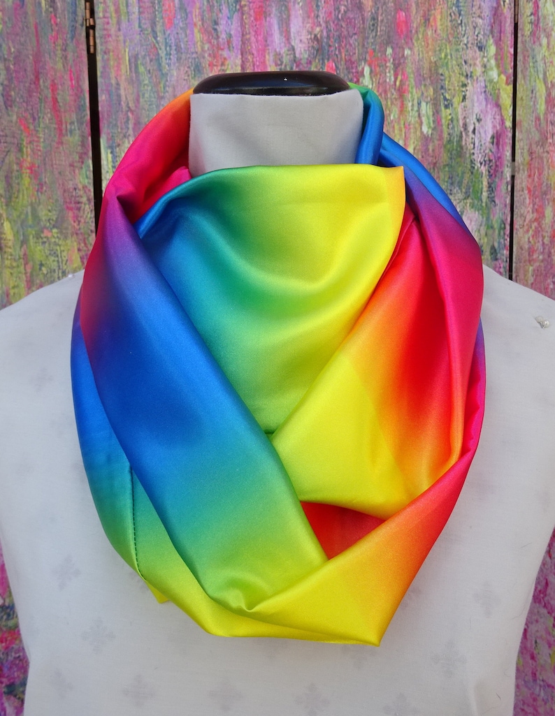 May include: A vibrant rainbow-coloured infinity scarf draped on a mannequin. The scarf displays a smooth gradient of colours, including red, orange, yellow, green, blue, and purple. The material appears to be a shiny, silky fabric.