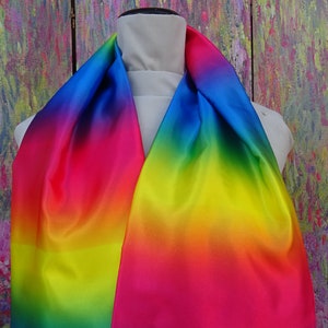 May include: A rainbow-coloured scarf on a mannequin. The scarf's colours blend from blue to pink, yellow, and green. The fabric has a glossy sheen, possibly satin or silk. The background is an abstract painting with a mix of colours.