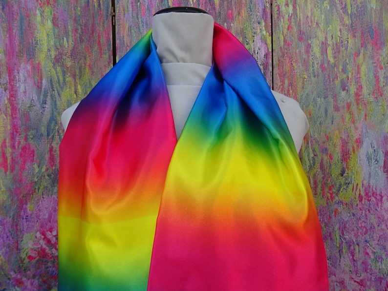 May include: A rainbow-coloured scarf on a mannequin. The scarf's colours blend from blue to pink, yellow, and green. The fabric has a glossy sheen, possibly satin or silk. The background is an abstract painting with a mix of colours.