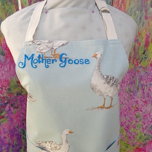 May include: Pale blue apron with goose illustrations and the embroidered text "Mother Goose" in blue. It has a light beige neck strap and a pocket. The background is a colorful floral design.