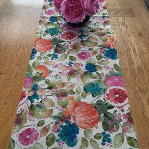 Puede incluir: Un camino de mesa floral con un dise&ntilde;o bot&aacute;nico con naranjas, pomelos y bayas azules. Un ramo de rosas rosas se encuentra encima del camino de mesa. El camino de mesa est&aacute; sobre una mesa de madera.