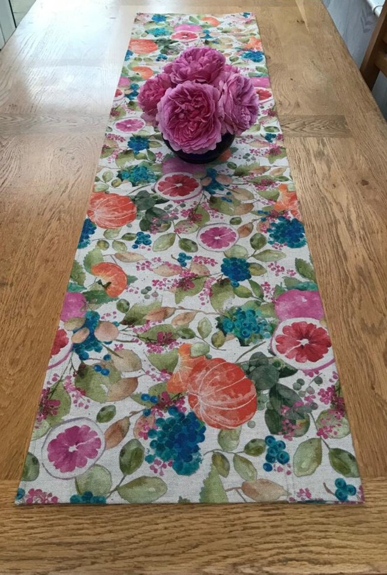 Puede incluir: Un camino de mesa floral con un dise&ntilde;o bot&aacute;nico con naranjas, pomelos y bayas azules. Un ramo de rosas rosas se encuentra encima del camino de mesa. El camino de mesa est&aacute; sobre una mesa de madera.