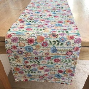 Può includere: Una tovaglia floreale con sfondo crema e un motivo ripetuto di fiori colorati nelle tonalità del rosa, blu, arancione e giallo. La tovaglia è rettangolare e realizzata in tessuto.