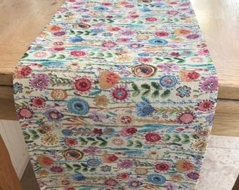 May include: A floral table runner with a cream background and a repeating pattern of colorful flowers in shades of pink, blue, orange, and yellow. The runner is rectangular and appears to be made of woven fabric.