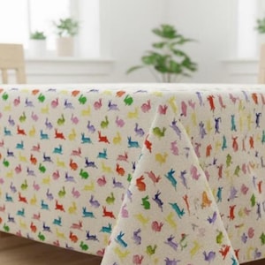 Könnte beinhalten: Ein rechteckiges Tischtuch in Creme mit einem sich wiederholenden Muster aus bunten Hasen in Rot, Orange, Gelb, Grün, Blau und Lila. Die Hasen sind in verschiedenen Posen, was ein verspieltes und festliches Design ergibt.