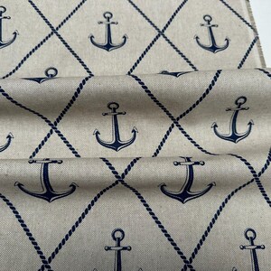 May include: A beige fabric featuring a repeating pattern of dark blue anchors centered within a diamond grid formed by dark blue rope-like lines. The material has a visible weave texture and frayed edges along the side of the folded textile.