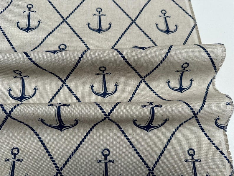 May include: A beige fabric featuring a repeating pattern of dark blue anchors centered within a diamond grid formed by dark blue rope-like lines. The material has a visible weave texture and frayed edges along the side of the folded textile.