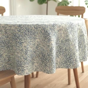 May include: A round tablecloth featuring a repeating botanical pattern of blue willow-like leaves on a cream-colored background. The fabric drapes over the edges of a circular table, displaying a dense, intricate foliage design across the surface.