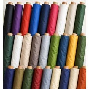 May include: A collection of fabric bolts arranged in rows, featuring a wide variety of solid colors including black, purple, red, blue, navy, pink, white, beige, green, gray, yellow, and orange, each with a visible wooden core at the top end.