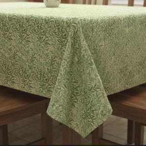 May include: A rectangular tablecloth featuring an all-over botanical print of overlapping willow leaves in various shades of green against a light cream background. The fabric drapes over the edges of a table, displaying a dense, repeating pattern.