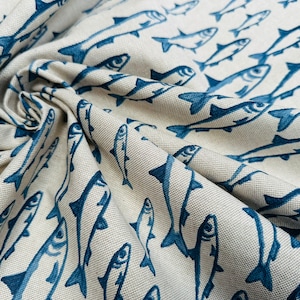 May include: A beige fabric featuring a repeating pattern of dark blue fish. The material has a visible woven texture and is gathered in folds, creating depth and shadows that highlight the orientation of the printed fish illustrations.