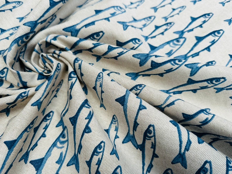 May include: A beige fabric featuring a repeating pattern of dark blue fish. The material has a visible woven texture and is gathered in folds, creating depth and shadows that highlight the orientation of the printed fish illustrations.
